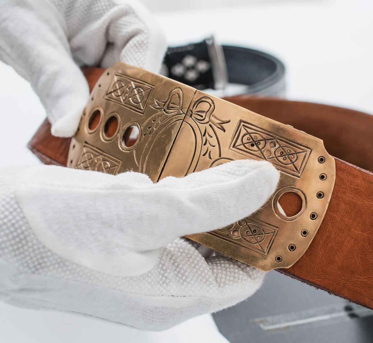 Zwei Hände mit weißen Handschuhen präsentieren die verzierte Bronzeschnalle eines mittelbraunen Ledergürtels.