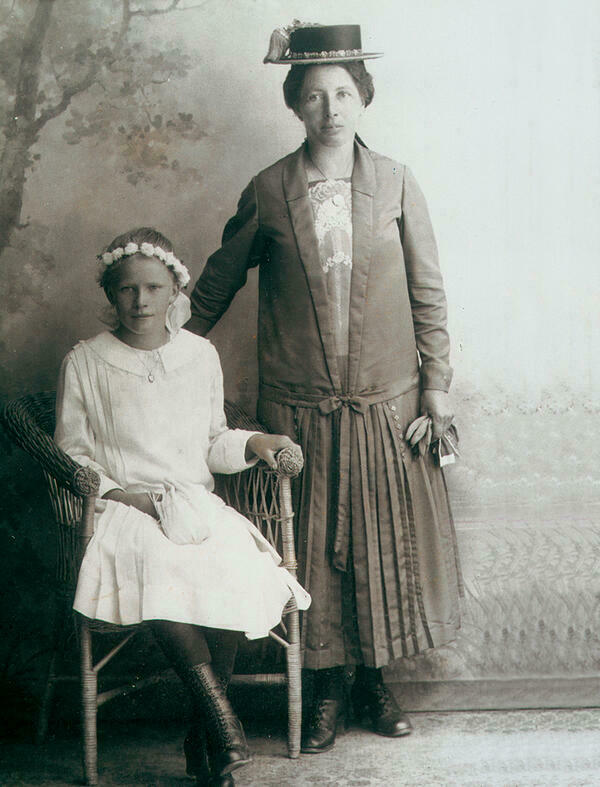 Die Schwarz-Wei-Photographie aus Attenberg um 1925/30 zeigt ein Mdchen in ihrem Firmkleid, das auf einem Korbstuhl sitzend neben ihrer festlich gekleideten Mutter in die Kamera lchelt. Hinter den beiden ist eine Landschaftstapete zu sehen.