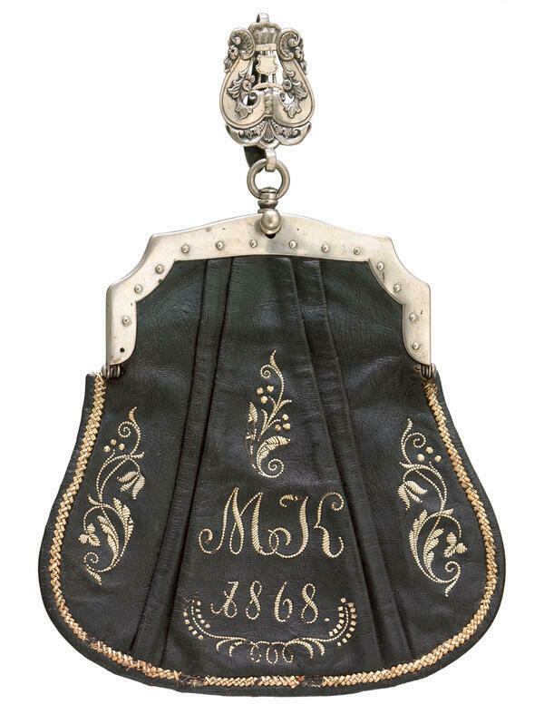 Die oberbayerische Kellnerinnen-Tasche aus dem Jahr 1868 ist aus schwarzem Leder gefertigt. Die Vorderseite zeigt ein Monogramm mit den Initialen MK, das ebenso wie die Jahreszahl und Schmuckmuster mit dem Federkiel aufgestickt wurden. Schliee und Aufhngung der Tasche sind aus versilbertem Messing.
