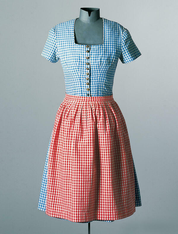 Die Kleiderpuppe trgt ein einfaches Baumwoll-Dirndl aus dem Raum Groweil um 1955. Das kurzrmlige Kleid ist blau-wei, die Schrze rot-wei kariert. Zehn Metallknpfe schlieen das Mieder.