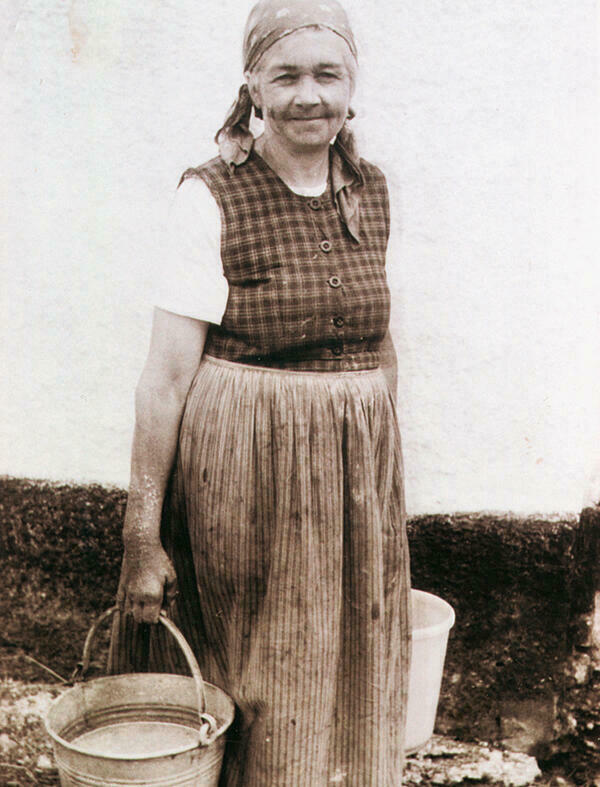 Die Schwarz-Wei-Photographie zeigt eine lchelnde ltere Buerin aus Rehling um 1950/60. Sie trgt ein gemustertes Arbeitskleid, darunter eine kurzrmelige weie Bluse, sowie ein Kopftuch. In beiden Hnden hlt sie jeweils einen Eimer.